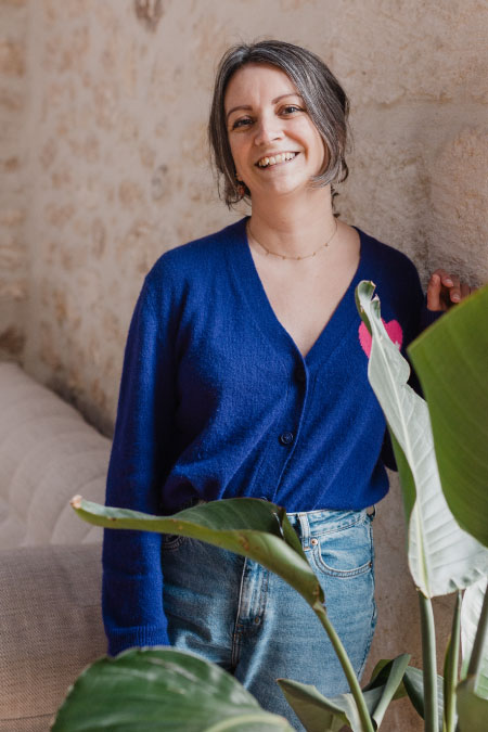 Steffi, créatrice de site Wordpress à Montpellier, sourit, appuyée contre un mur de pierre, avec une plante à l'avant plan